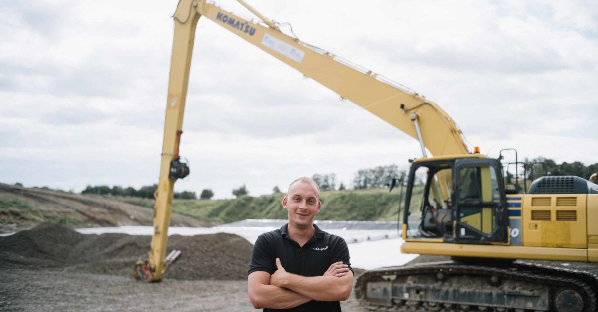 Portrait eines Mitarbeiters mit kurzen blonden Haaren. Er verschränkt die Arme und schaut selbstsicher in die Kamera. Im Hintergrund ist ein gelber Bagger zu sehen.