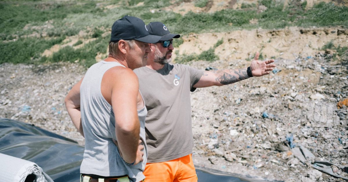 Zwei Kollegen stehen auf einer Deponie und schauen in die Ferne. Sie tragen beide Firmen- und Sicherheitskleidung, der eine Kollege zeigt auf etwas.