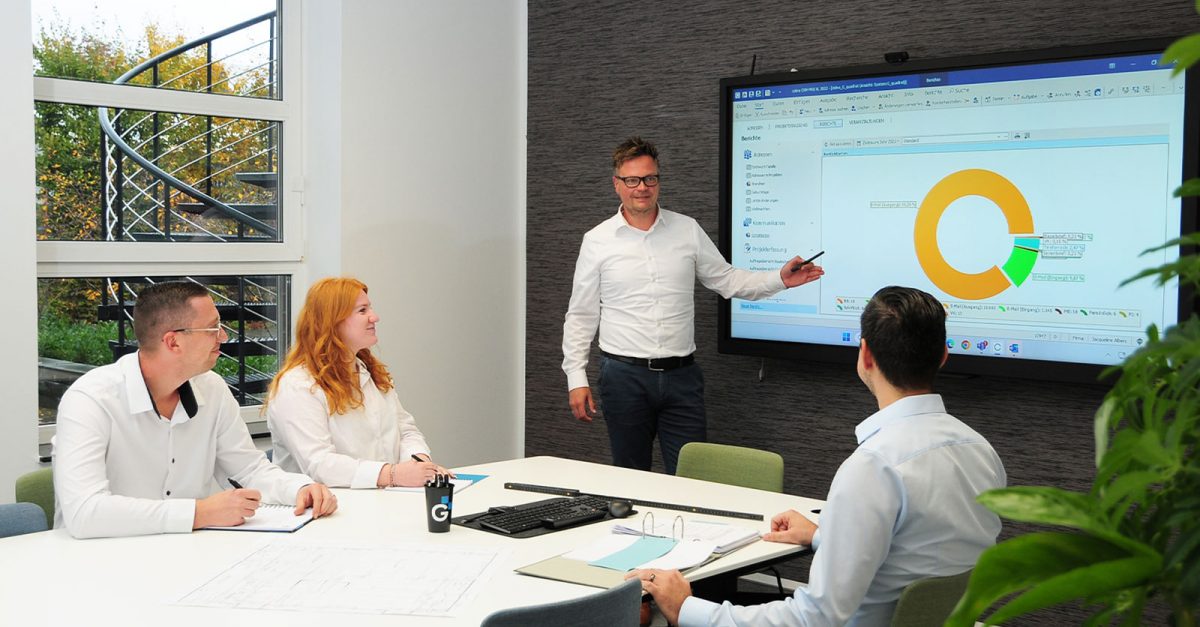 Besprechungsraum, ein Kollege präsentiert ein Diagramm an einem Smartboard, 3 weitere Kollegen sitzen an einem Tisch und hören aufmerksam zu.