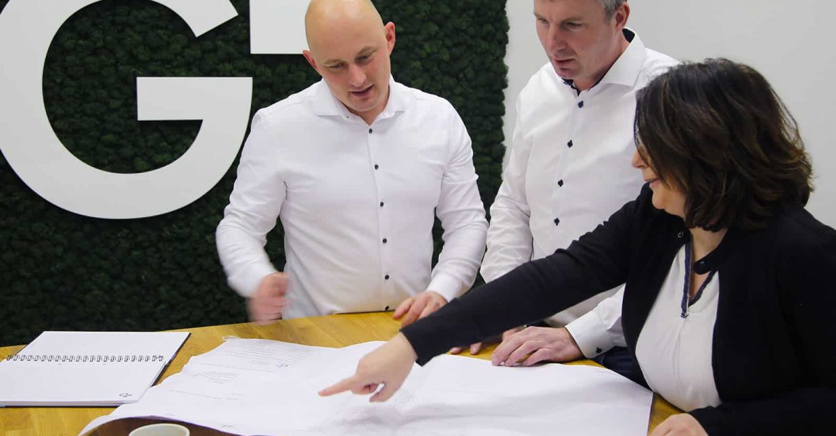 3 Kolleg:in stehen im Halbkreis vor dem Firmenlogo an einem Stehschreibtisch und blicken auf einen Plan. Eine Kollegin zeigt mit ihrem Finger auf den Plan.